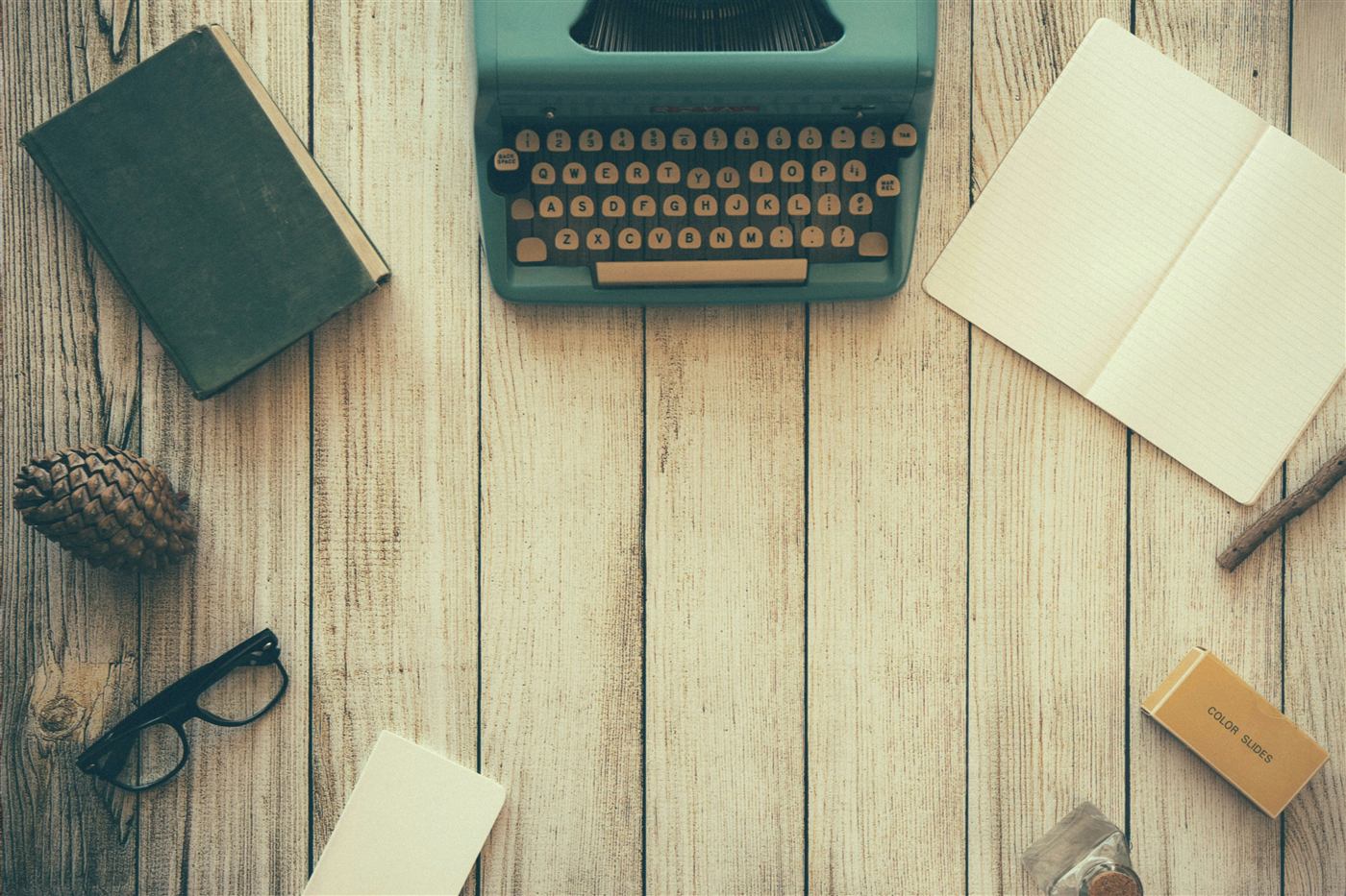 Vintage typewriter, notebook, book, glasses, and pine cone arranged on a wooden desk